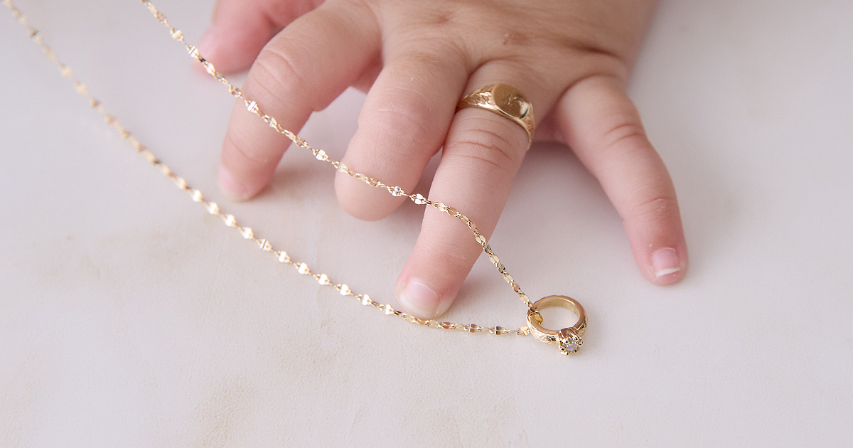 赤ちゃんの手に着けたイニシャルシグネットベビーリングと、隣に置かれたチェーン付きバースデーストーンベビーリング